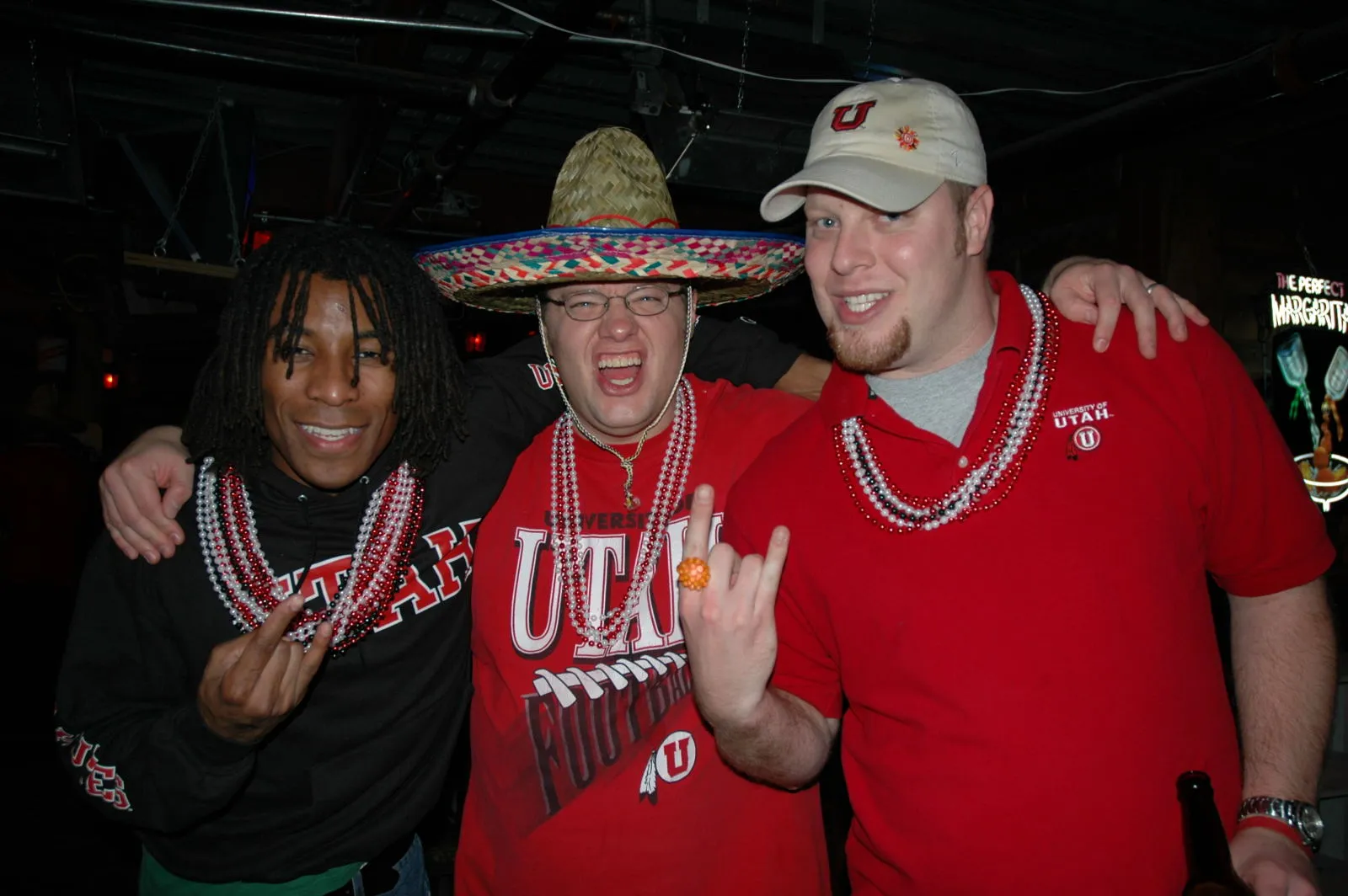 Three friends in Utah gear and beads, one in a sombrero, at a night tailgate
