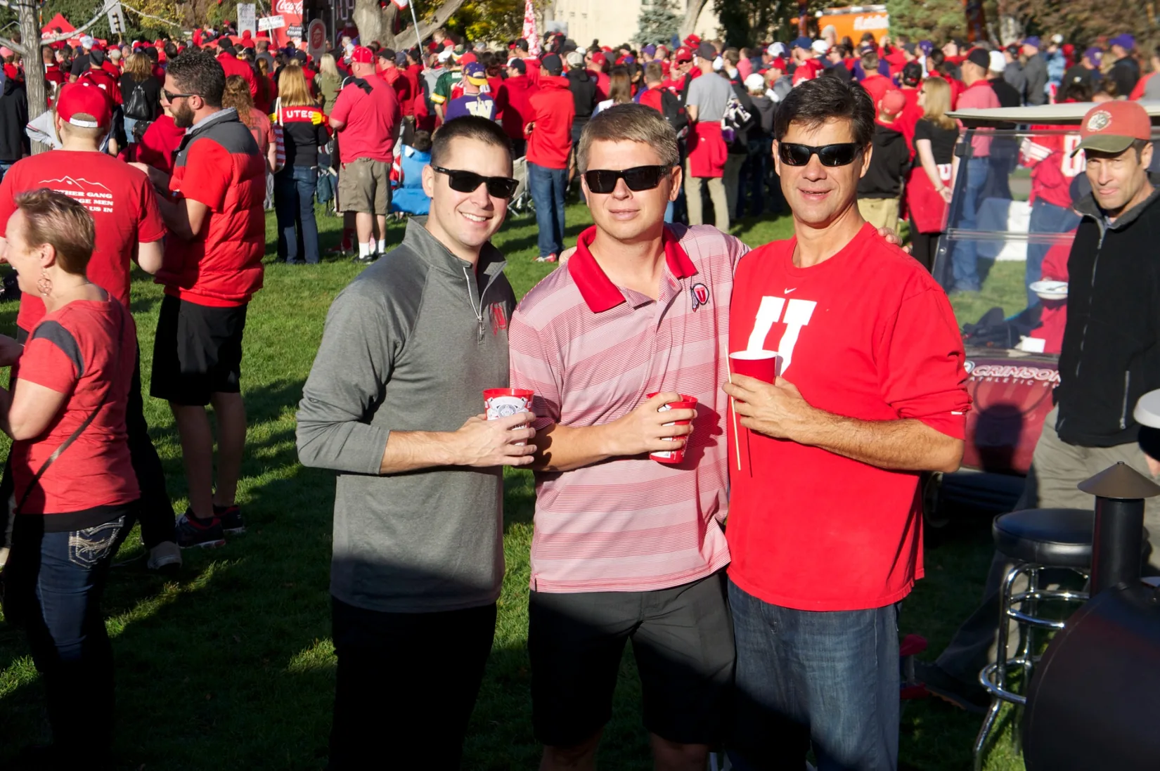 Three friends in Utah gear holding red cups in a crowded tailgate field