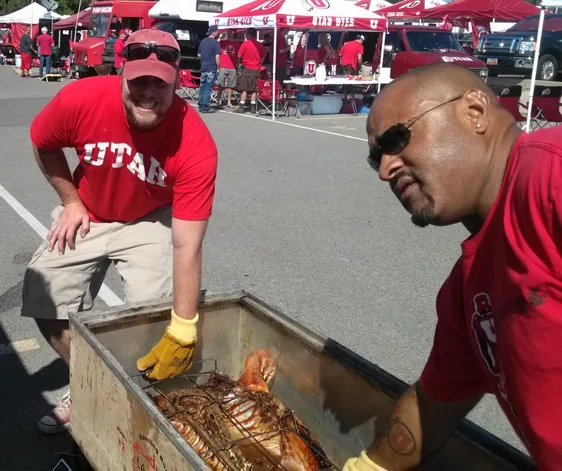 A crew member in a Utah shirt and gloves tending a whole pig on the roaster